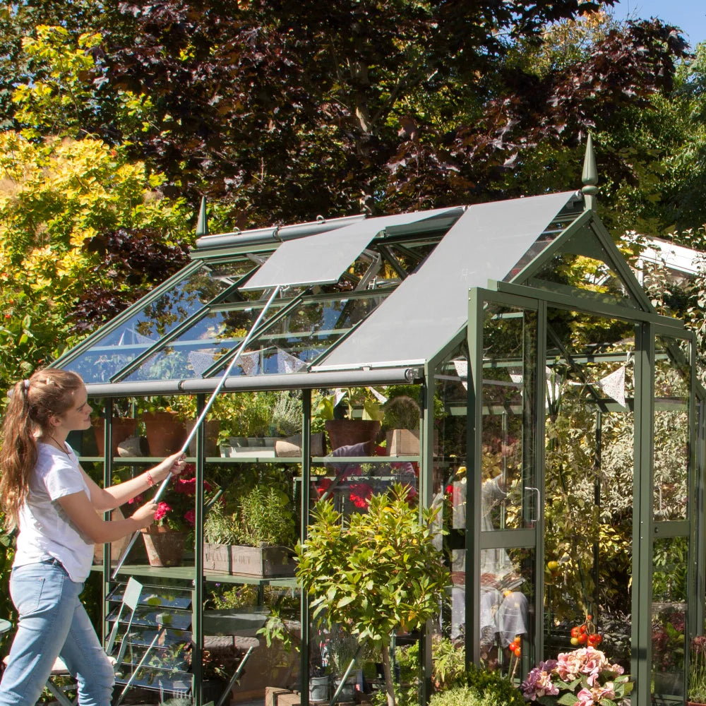 A Rhino Greenhouse with open roof panels is being adjusted by a person using a pole. Inside, there are potted plants and gardening accessories. Surrounding the structure are lush trees and shrubs.
