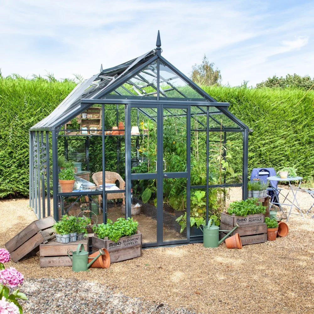 A glass Rhino Greenhouse containing various plants and a wicker chair stands on a gravel path surrounded by green hedges. Nearby, crates with plants and watering cans are placed. Text on the crates: Rhino Norfolk and Bloomin' Lovely!.