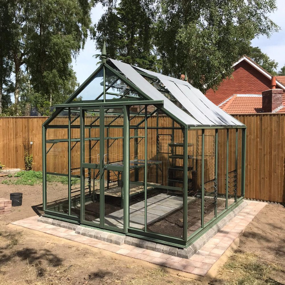 A green-framed glass Rhino Greenhouse stands on a paved base, surrounded by a wooden fence and trees. In the background, red-roofed houses are partly visible.