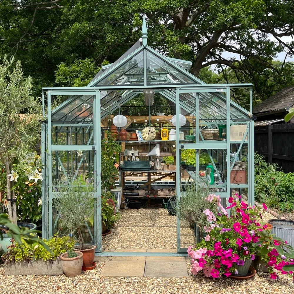 A mint-colored Rhino Greenhouse is filled with plants and gardening tools; it sits on a gravel path surrounded by vibrant potted flowers and leafy trees in a lush garden setting.
