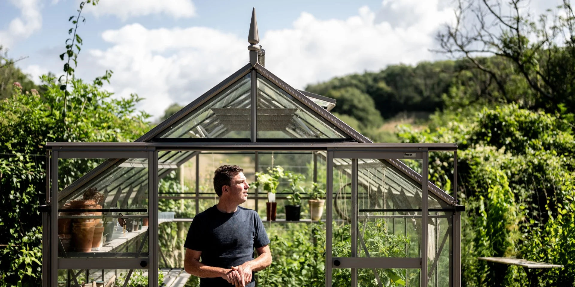 A person stands in front of a glass Rhino Greenhouse, surrounded by lush greenery under a partly cloudy sky. The Rhino Greenhouse is filled with plants and gardening tools.