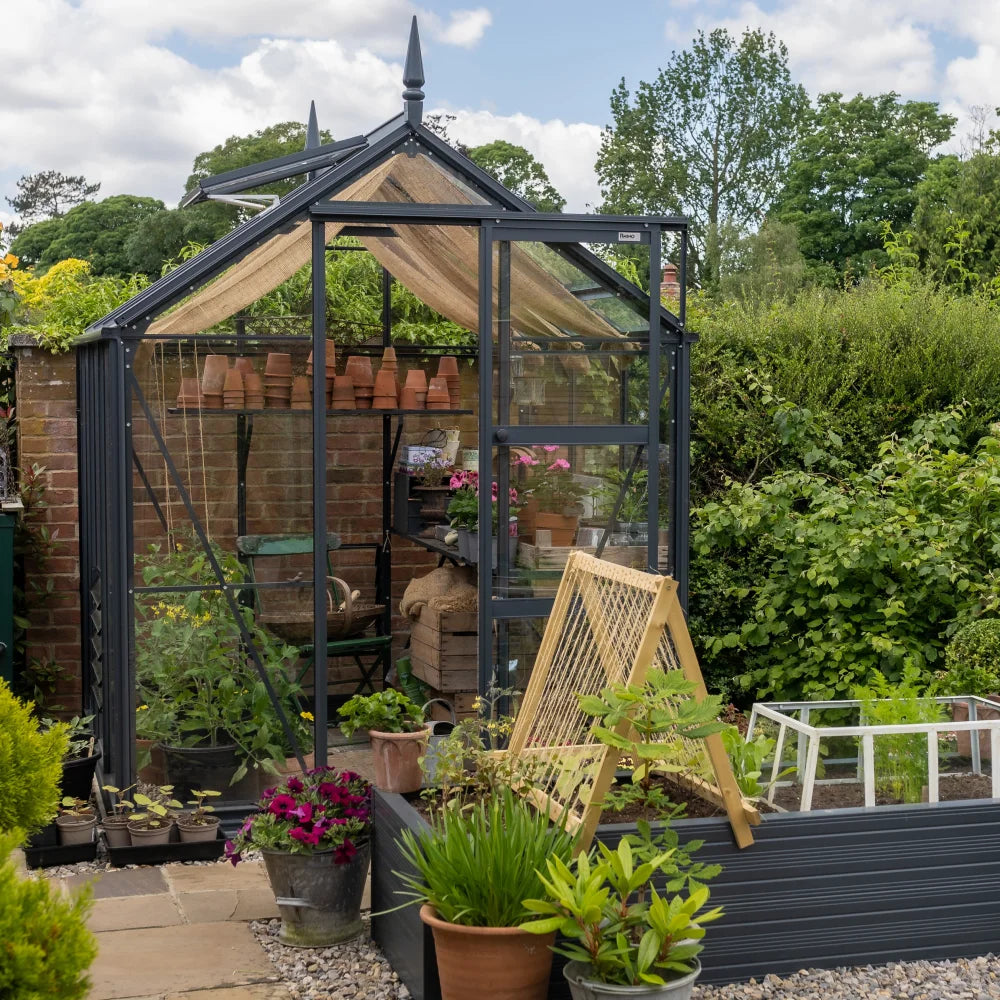 A glass Rhino Greenhouse contains various potted plants and gardening tools, with additional plants and a small trellis outside. It is set against a lush garden backdrop with trees and hedges.