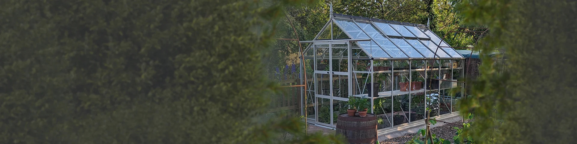 A glass-paneled Rhino Greenhouse filled with potted plants and greenery stands in a garden area, surrounded by trees and foliage, with a wooden barrel and path nearby.