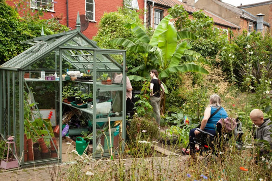 A glass Rhino Greenhouse filled with plants and gardening tools stands in a lush garden. A person in a wheelchair and a child engage with the surroundings, near a large banana plant.