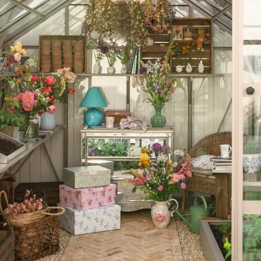 The Rhino Greenhouse interior features various colorful flowers in vases and baskets, a mirrored table with a turquoise lamp, and decorative boxes. A wicker chair and gardening tools enhance the cozy atmosphere.