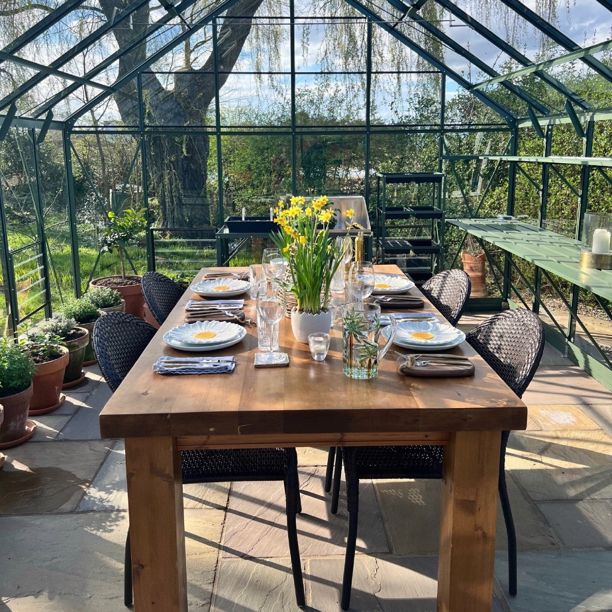 A wooden table set for dining, featuring a vase of yellow flowers, stands inside a glass Rhino Greenhouse. Surrounding the table are potted plants and garden scenery outside.