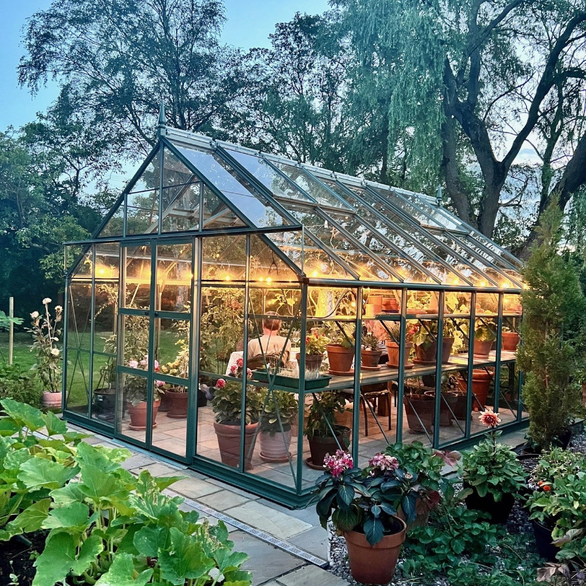 A Rhino Greenhouse filled with potted plants is illuminated warmly inside a lush garden. A person tends to the plants, surrounded by mature trees and a clear evening sky.