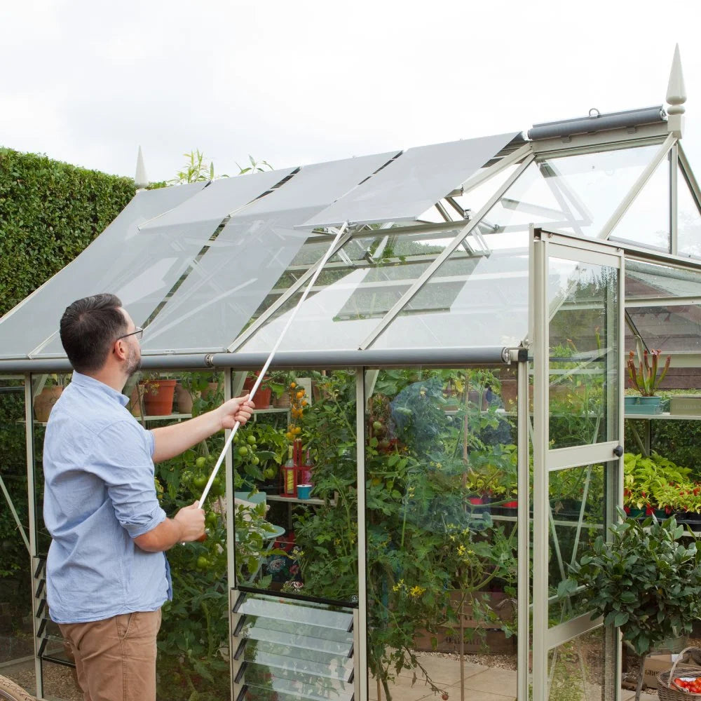 A person adjusts a retractable shade over a glass Rhino Greenhouse using a pole, surrounded by lush plants and a neatly trimmed hedge in a garden setting.