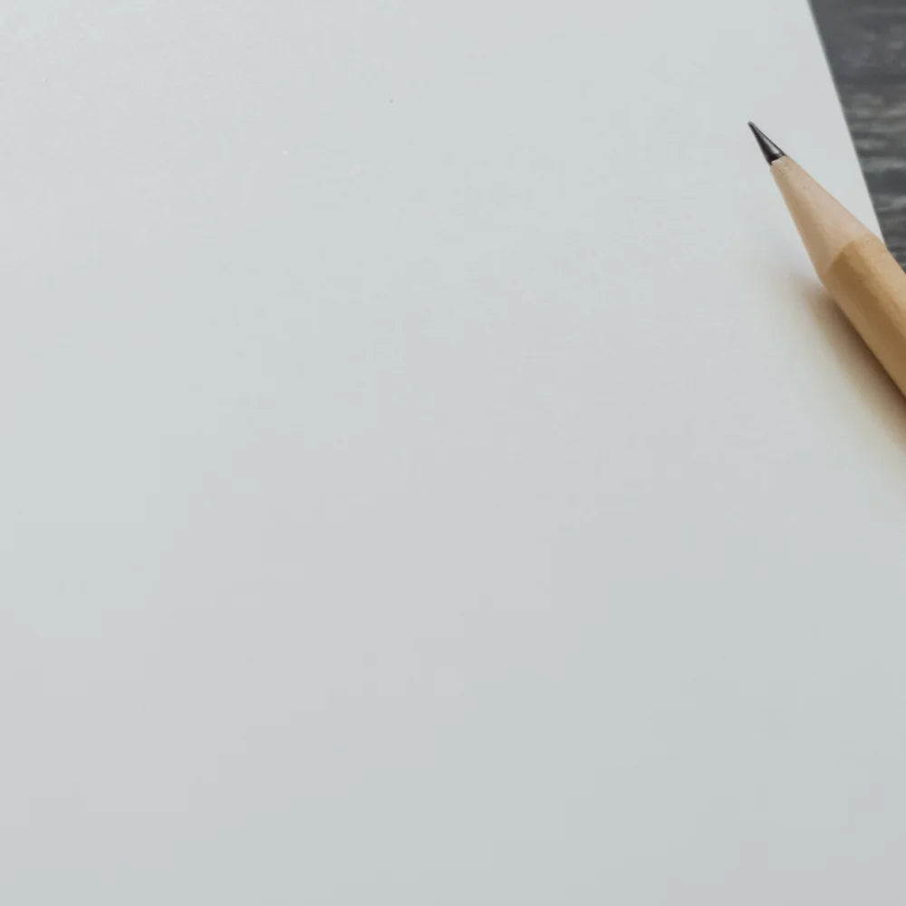 A sharpened pencil rests on a blank white sheet of paper, lying on a textured dark surface, suggesting a writing or sketching context.