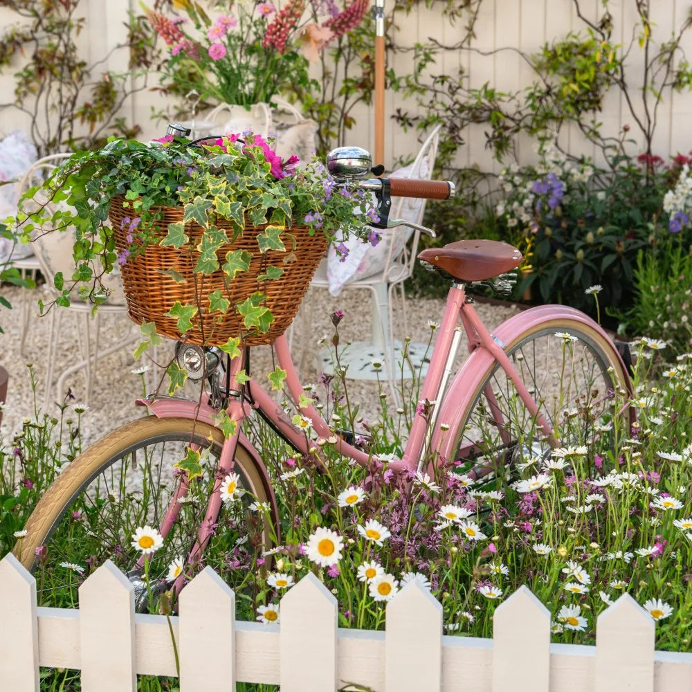 A vintage pink bicycle with a wicker basket overflows with greenery and flowers, parked in a lush garden behind a white picket fence, surrounded by colorful blooming plants and chairs.