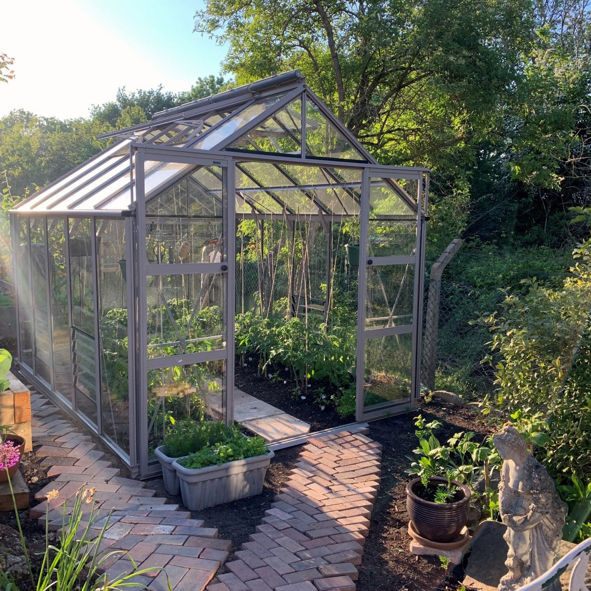 A glass Rhino Greenhouse stands in a sunlit garden, housing thriving plants inside. A brick pathway leads to it, surrounded by greenery and a small garden statue.