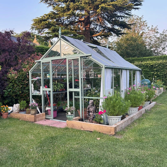 A green Rhino Greenhouse with glass panels houses various plants inside, surrounded by potted flowers and wooden planters, set in a lush garden with tall trees in the background.