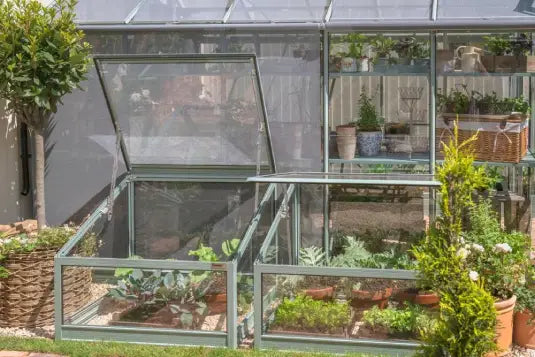 Greenhouses with open vents house various plants in pots. Surrounding the structures are tall greenery and additional potted plants, set in a garden landscape.