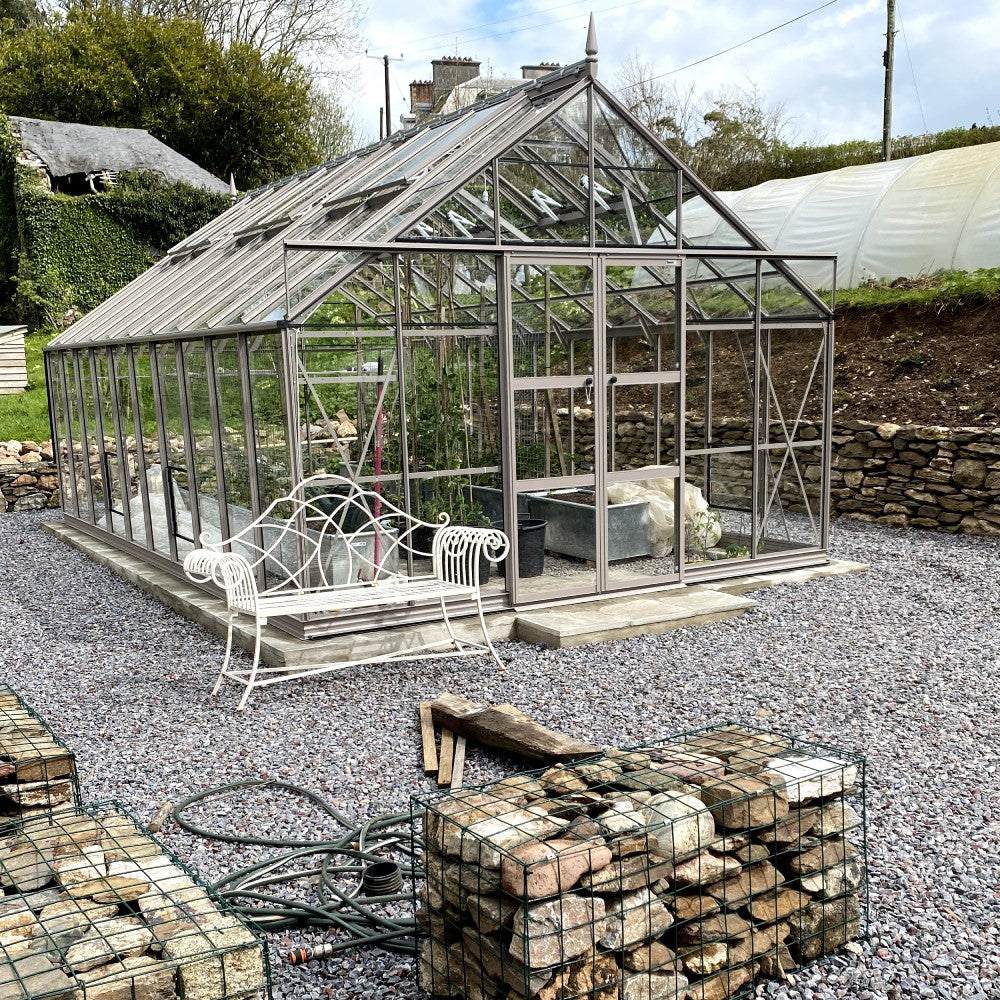 A metal-framed Rhino Greenhouse with glass panels stands amidst gravel, housing plants inside. A decorative white bench sits nearby, surrounded by wired rock cages and gardening tools in a garden setting.