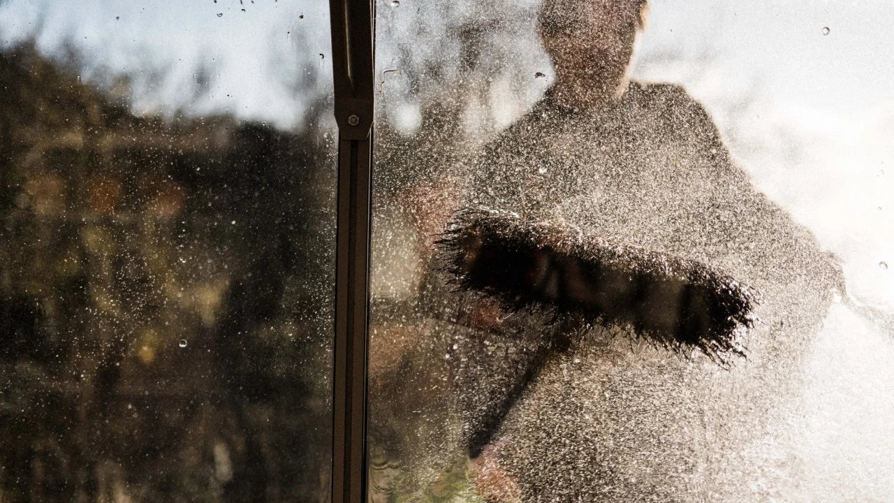 A person is cleaning a glass surface with a brush, creating a splash of water droplets, while sunlight filters through trees in the background.