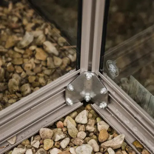 A metal bracket secures aluminum Rhino Greenhouse frames, forming a sturdy corner, resting on a bed of small pebbles.