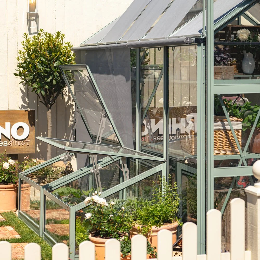 Rhino Greenhouse with open windows, framed by potted plants, stands in a garden. A sign reads RHINO Rhino Rhino Greenhousesdirect on the side wall. A white picket fence surrounds the area.