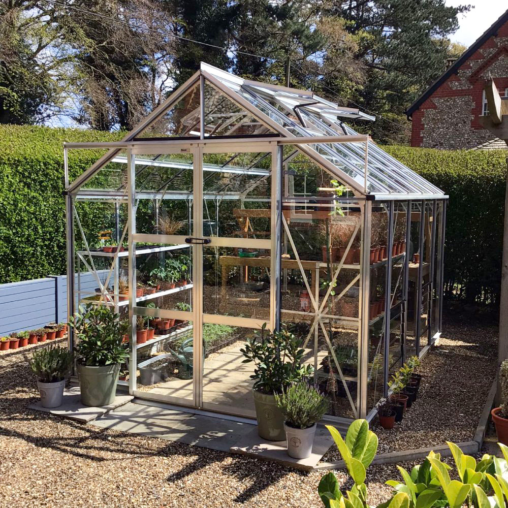 A glass Rhino Greenhouse filled with plants stands on a gravel surface, surrounded by potted plants and hedges, next to a brick building. The scene is outdoors with trees in the background.
