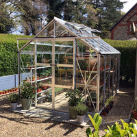 A glass Rhino Greenhouse filled with plants stands on a gravel surface, surrounded by potted plants and hedges, next to a brick building. The scene is outdoors with trees in the background.