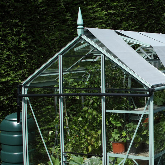 A glass Rhino Greenhouse with triangular roof panels shelters lush green plants and potted greenery. It is set within a dense hedge-lined garden, suggesting a focus on outdoor gardening.