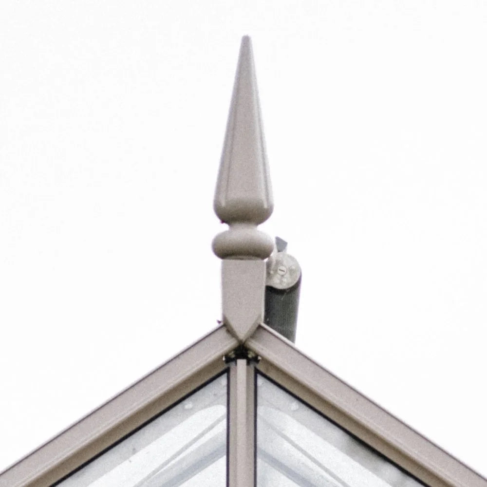 A pointed metal finial tops the intersecting edges of a glass Rhino Greenhouse roof. The finial is prominently displayed against a plain white background, emphasizing architectural detail.