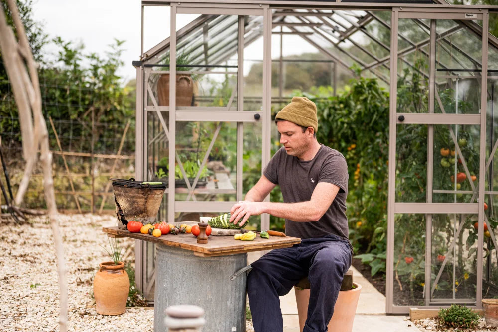 A person slices vegetables on a table outside a Rhino Greenhouse filled with plants. Surrounding the area are various gardening supplies and lush greenery, creating a vibrant, productive garden setting.