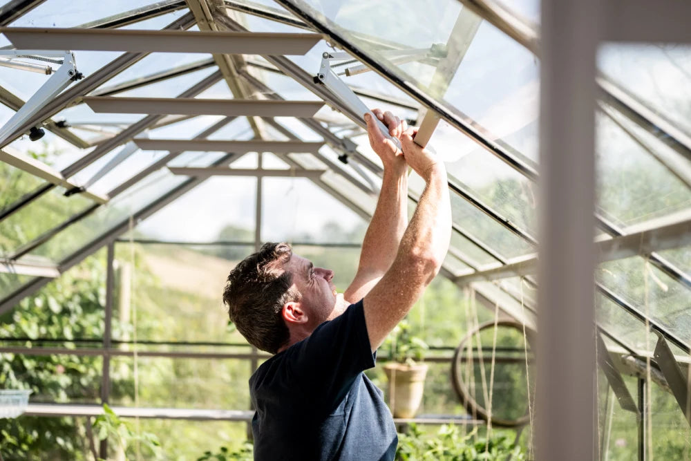 A person adjusts a window panel inside a Rhino Greenhouse. Sunlight streams through the glass roof, surrounded by lush green plants outside and hanging pots within.