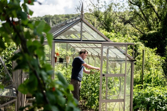 A person adjusts the door of a glass Rhino Greenhouse surrounded by lush plants. Nearby, potted plants rest on shelves inside, and dense greenery surrounds the structure under a clear sky.