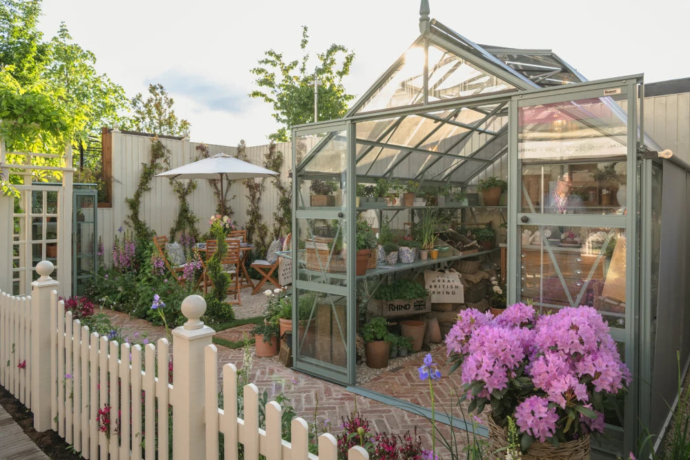 Rhino Greenhouse with open doors showcases potted plants on shelves. A small garden with blooming flowers and outdoor seating lies beyond a white picket fence. Text reads The Great British Rhino.