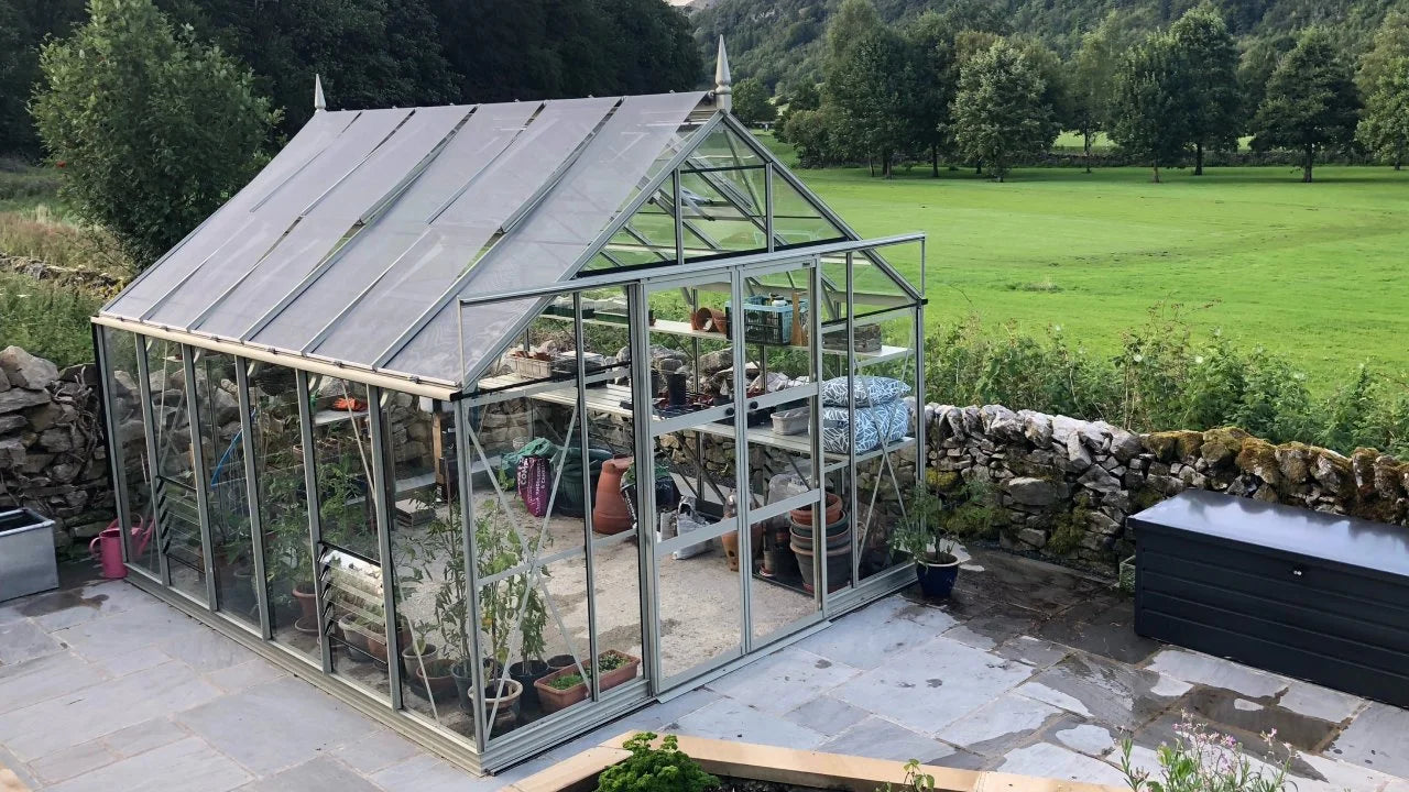 A glass Rhino Greenhouse shelters various gardening tools and potted plants, situated on a stone patio beside a stone wall, with a lush green landscape in the background.