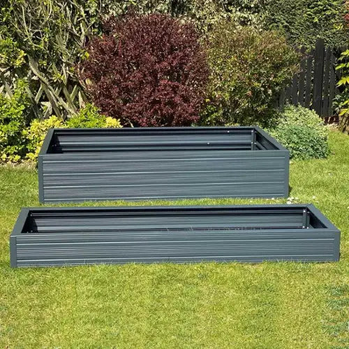 Two rectangular, dark gray Rhino metal raised garden beds are positioned on a green lawn. The background features a variety of shrubs and plants, creating a lush garden setting.