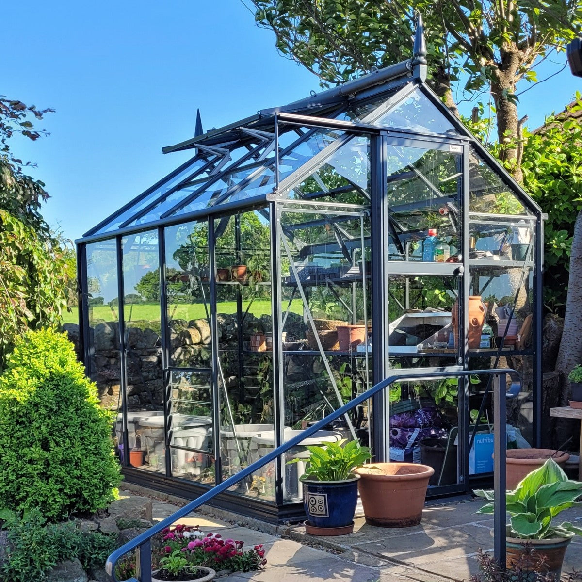 A glass Rhino Greenhouse stands in a garden, filled with potted plants on shelves. A stone wall and trees surround it, with additional potted plants placed along a stone pathway.
