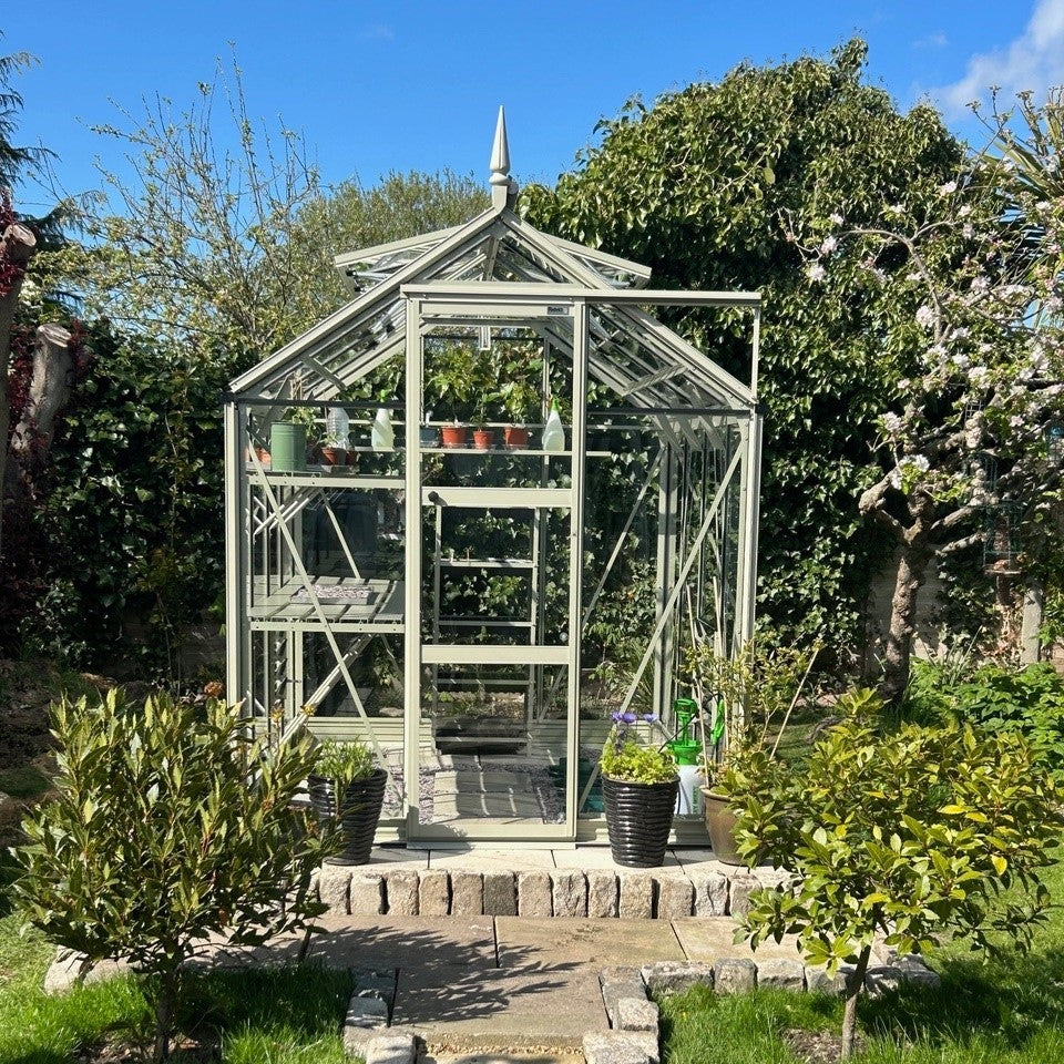 A glass Rhino Greenhouse houses various potted plants on shelves, surrounded by a lush garden with trees and shrubs under a clear blue sky.