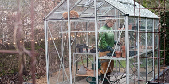 A person tends to plants inside a glass Rhino Greenhouse, surrounded by gardening tools. Outside, string lights hang against a backdrop of trees and a stone wall, adding charm to the scene.