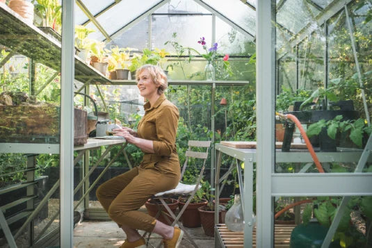 A person sits on a folding chair inside a glass Rhino Greenhouse, tending to potted plants on metal shelves. Sunlight filters through the glass, illuminating the lush greenery.