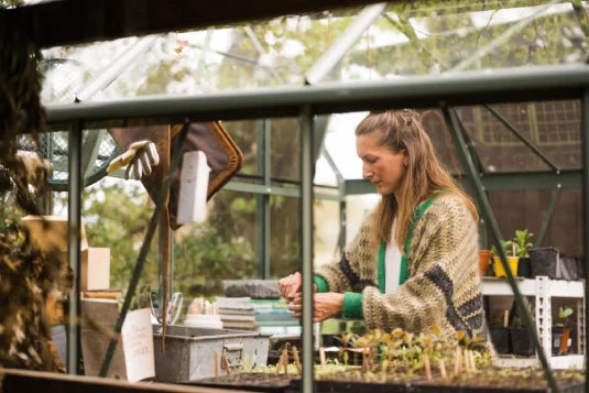Woman tending plants inside a Rhino Greenhouse, surrounded by gardening tools and seed trays. She wears a patterned sweater. Outside, leafy surroundings are visible through the glass panes.