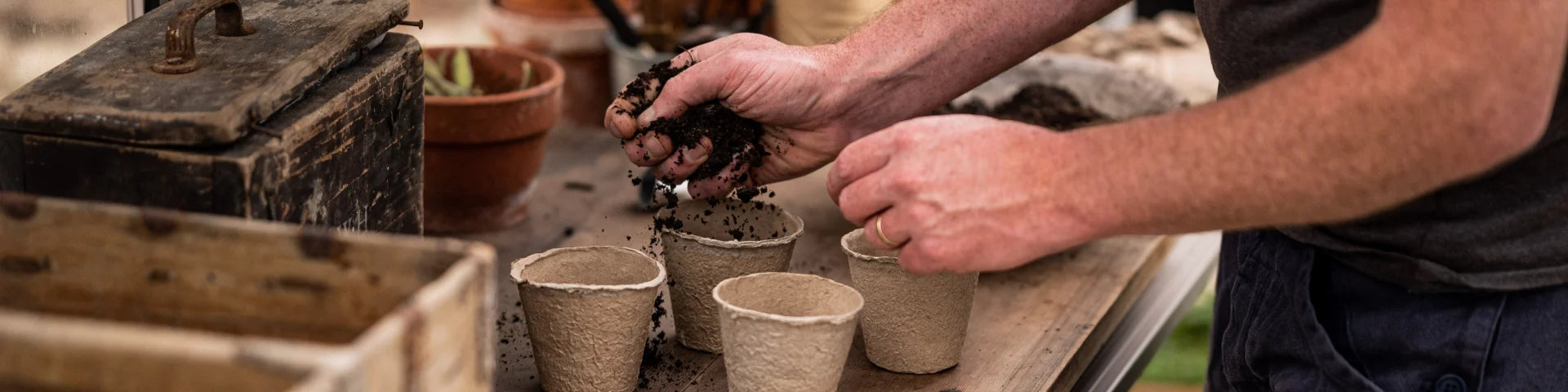 Hands are filling biodegradable pots with soil. They are on a wooden table surrounded by gardening tools and a potted plant, inside a Rhino Greenhouse setting.