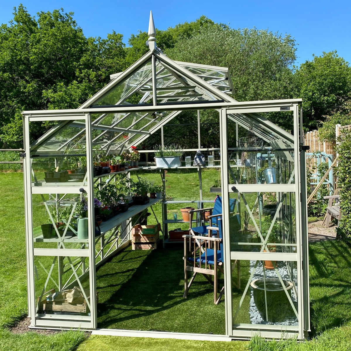 A glass Rhino Greenhouse stands open, showcasing rows of potted plants and gardening tools inside. It is set against a lush garden with trees under a clear blue sky.