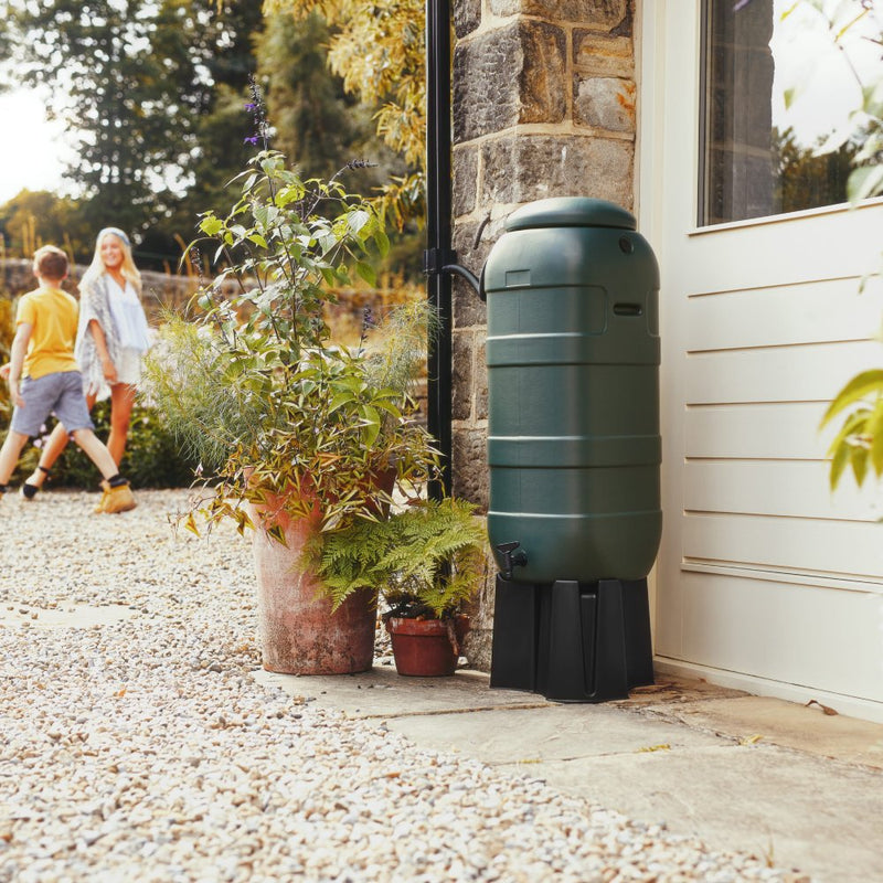 A green rain barrel stands beside a stone house, surrounded by potted plants. In the background, a child and a woman walk on a gravel path.