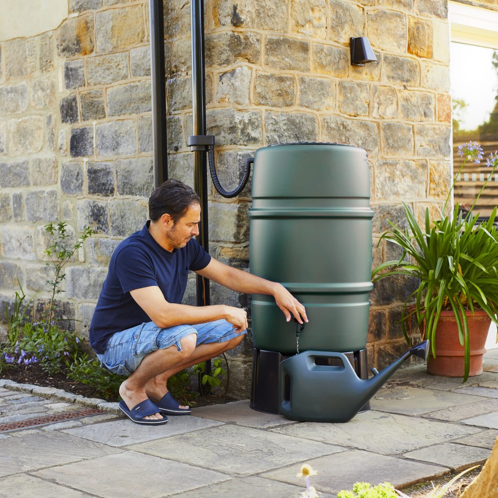 A man fills a watering can from a large green rain barrel. He is crouching on a stone patio beside a stone building with plants in the background.