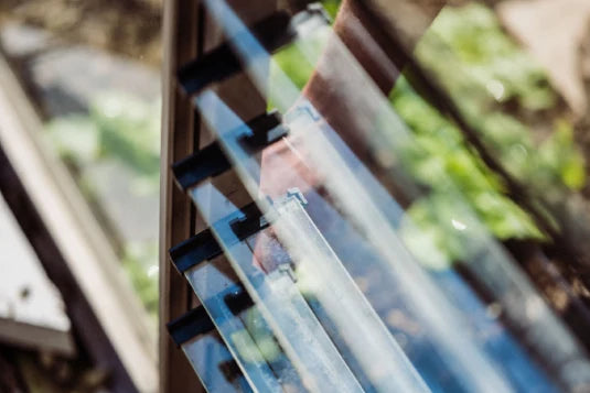 Glass Rhino Greenhouse panels partially open, reflecting light. A hand adjusts the panels. In the background, blurred greenery can be seen, suggesting a garden setting.