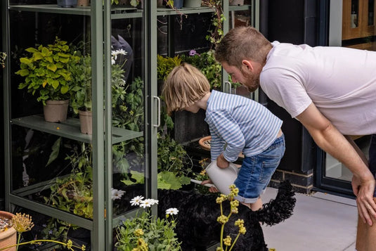 A child waters plants inside a green Rhino Greenhouse, assisted by an adult. Nearby, a black dog sits on a patio surrounded by potted flowers and greenery.