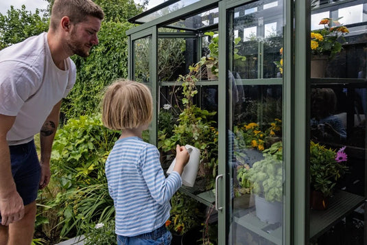 Child waters plants inside a glass Rhino Greenhouse, observed by an adult. Various green plants and colorful flowers fill the shelves. Lush garden surrounds them under a cloudy sky.