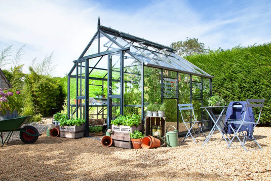 A glass Rhino Greenhouse stands in a garden, surrounded by potted plants and gardening tools. Nearby, blue chairs and a table sit on a gravel path. Text on crates reads Blooming Lovely and Rhino, handbuilt in Norfolk.