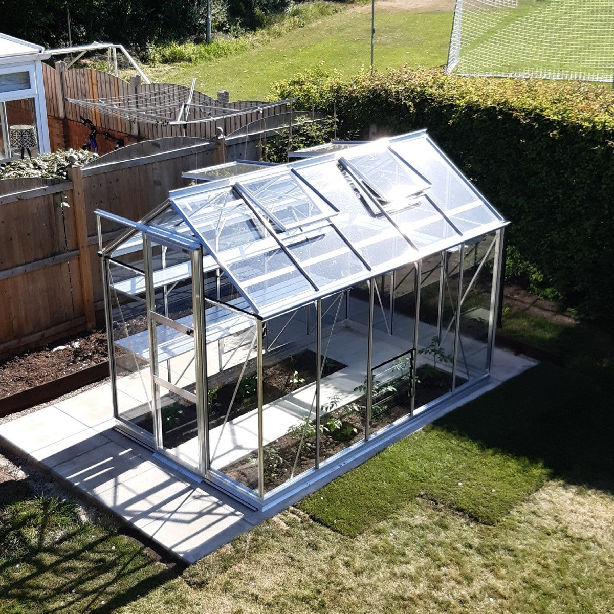 A glass-paneled Rhino Greenhouse with open roof vents sits on a concrete base, surrounded by a grassy yard and wooden fencing. Inside, plants are growing along the soil beds.