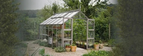 A glass Rhino Greenhouse stands surrounded by potted plants and gravel paths, nestled in a lush, green rural landscape with trees and hills in the background.