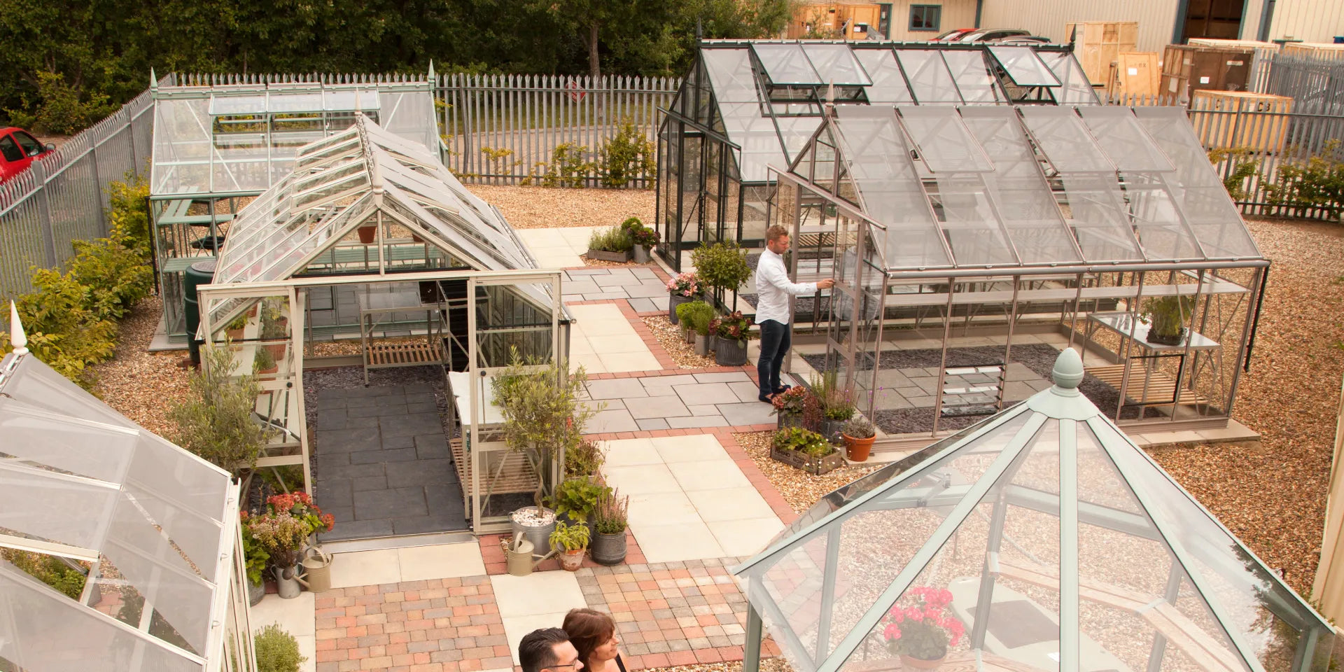 Greenhouses with clear panels are displayed in a garden center, surrounded by potted plants and paved paths; a person examines one structure.