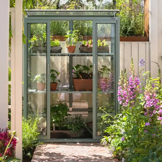 A glass Rhino Greenhouse houses potted plants on shelves, surrounded by lush flowering plants in a sunlit garden.
