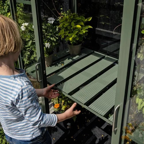 A child opens a Rhino Greenhouse door, revealing potted plants on shelves inside. The setting is lush, with more plants visible in the background, suggesting a garden environment.