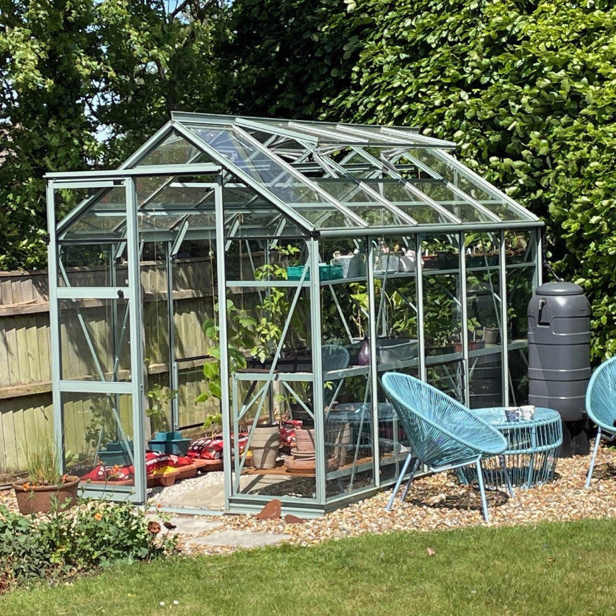 A glass Rhino Greenhouse stands in a garden surrounded by lush greenery. Inside, plants are visible. Blue chairs and a table are positioned outside on a pebble surface.